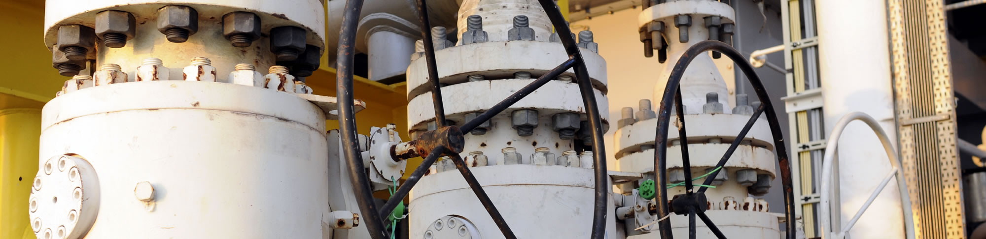 A close-up, panoramic view of large, industrial-looking white pipes and flanges, with black manual handwheels and numerous nuts and bolts visible, suggesting a complex valve system or a control panel.