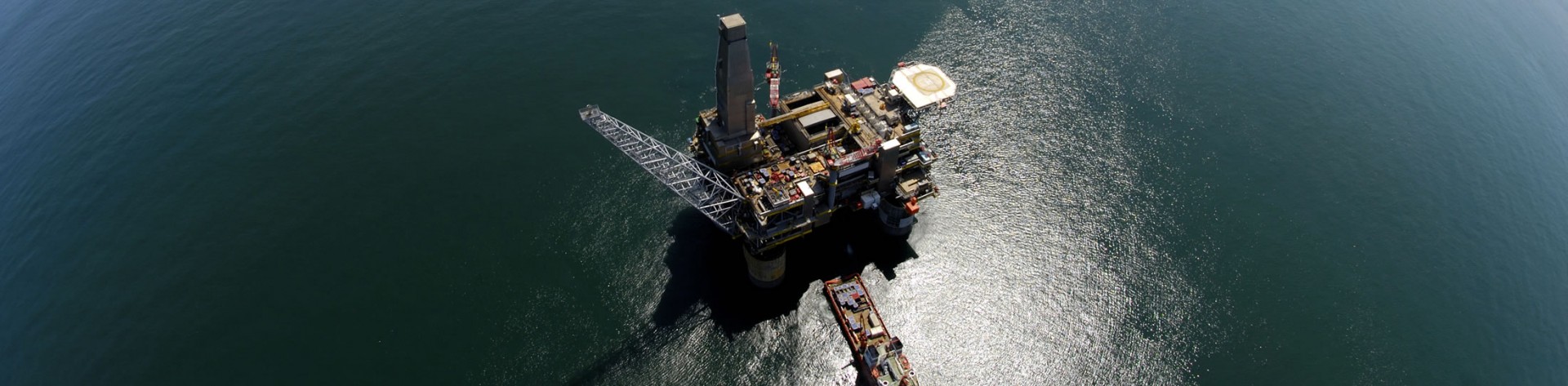 A top-down aerial view of a large offshore oil platform and an attached supply vessel floating in the deep blue ocean. The platform casts a long shadow on the water.