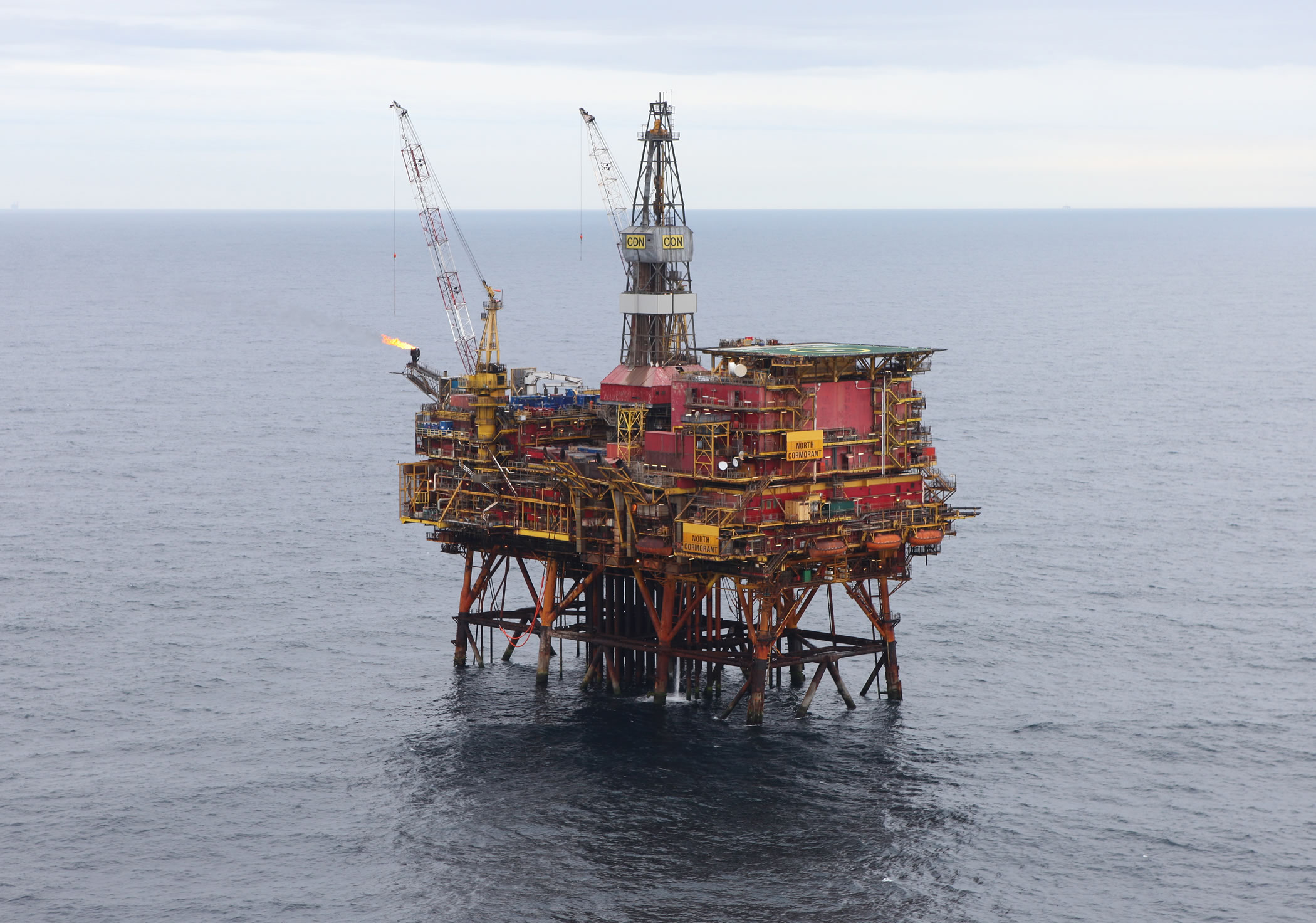 An aerial view of the North Cormorant oil platform, a large red and yellow structure with two cranes, standing in the middle of a calm, gray sea under a cloudy sky.