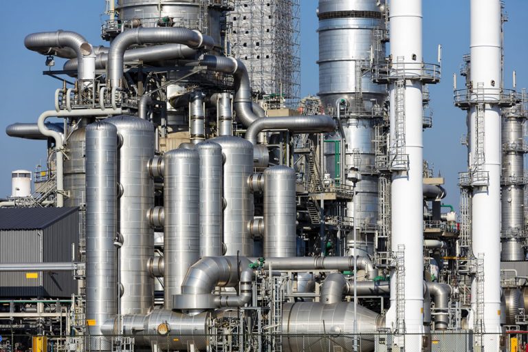 Industrial refinery with large silver metal pipes, tanks, and towers interconnected under a clear blue sky.