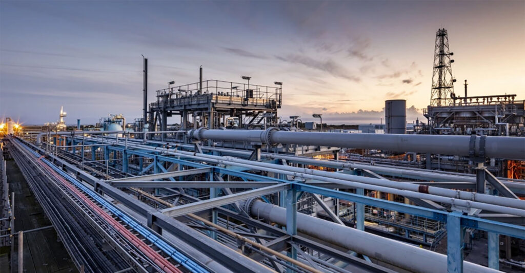 Industrial facility with large metal pipelines, structural frameworks, and processing equipment at sunset. The scene features complex piping and machinery typical of a refinery or plant.