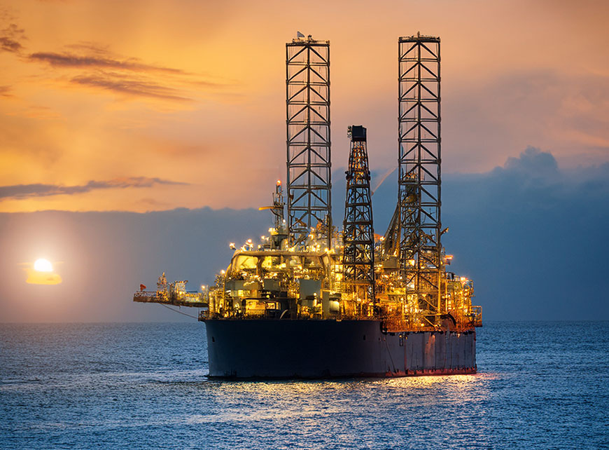 Offshore oil rig illuminated at sunset, positioned in calm ocean waters with a glowing sky and sun on the horizon.