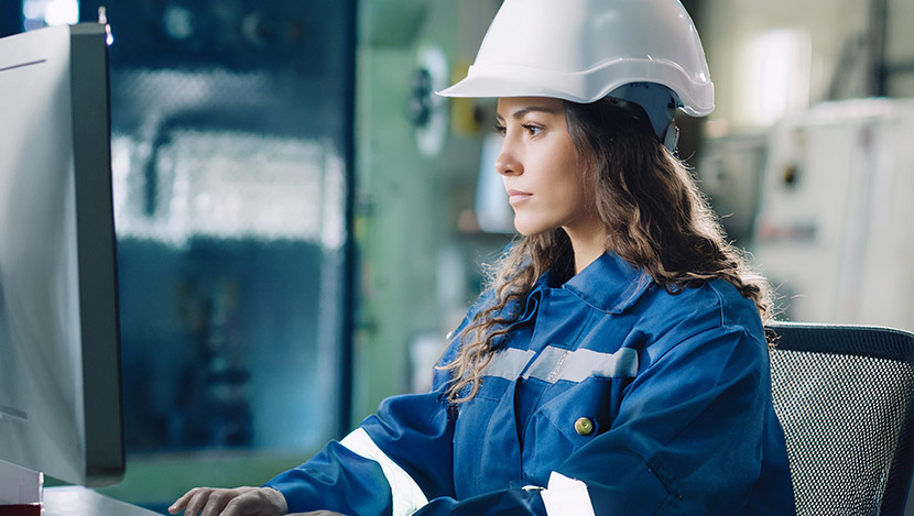 A woman wearing a hard hat and blue safety uniform works at a computer in an industrial or factory setting.