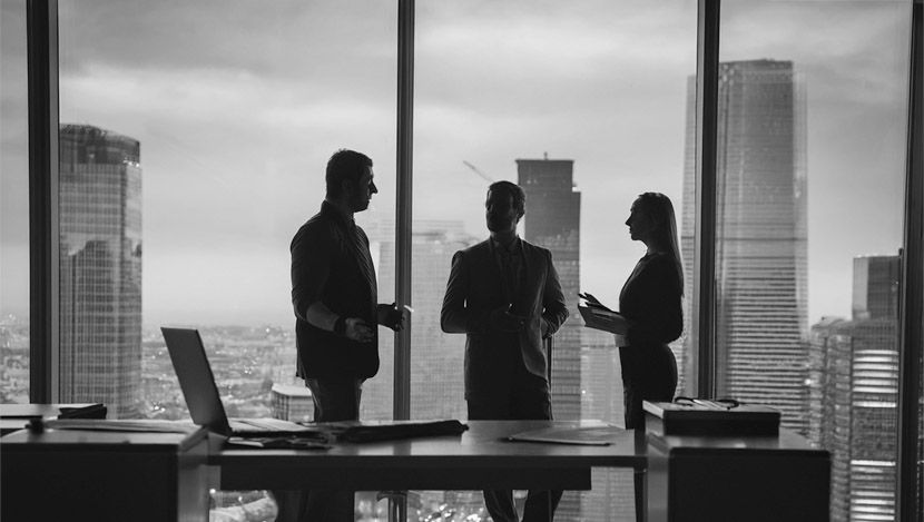 Three people in business attire stand and converse in an office with large windows, overlooking a city skyline with tall buildings.